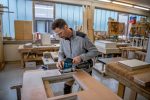 Eine Person mit Schutzausrüstung schleift in einer Werkstatt Holz mit einem Schwingschleifer.